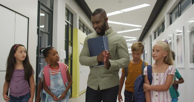 Teacher is leading a group of diverse students through a school corridor, creating a sense of unity and teamwork. The children, who are carrying backpacks, appear to be having an animated discussion with their teacher, emphasizing a positive learning environment. This image is perfect for illustrating concepts related to education, childhood, diversity, mentorship, and school life.