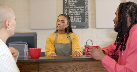 Female barista engaging customers at cozy coffeehouse counter with red mugs and chalkboard