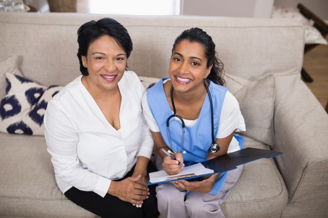 Smiling patient sitting with doctor reviewing medical report on sofa at home. Ideal for illustrating home healthcare services, elderly care, medical consultations, and patient-doctor relationships. Useful for healthcare websites, brochures, and advertisements promoting home medical visits and personalized care.
