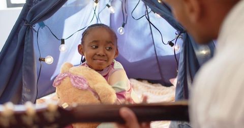 African american girl listening to guitar in cozy tent holding teddy under string lights