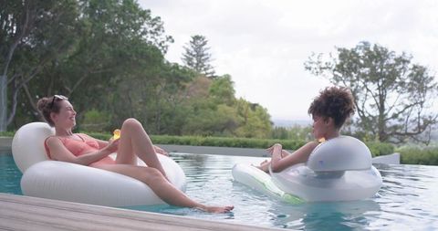 Diverse women relaxing on white inflatable floats in backyard pool enjoying summer leisure