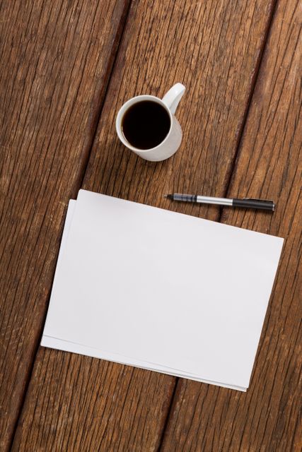 Cup of coffee next to blank sheet of paper and pen on wooden table. Ideal for illustrating concepts of planning, creativity, brainstorming, or morning routines. Suitable for use in articles, blogs, or advertisements related to workspaces, office environments, or productivity.