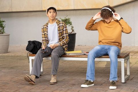 Two male students sitting on campus bench, one with smartphone, other adjusting headphones