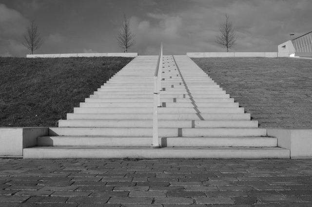 Concrete staircase positioned symmetrically between grassy slopes and paved area. Ideal for use in themes related to urban design, architecture, minimalism, and monochromatic art.