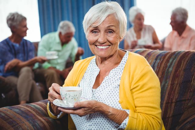 Senior woman smiling while holding a cup of coffee, with friends socializing in the background. Ideal for use in advertisements for retirement communities, senior living facilities, coffee brands, or social clubs. Highlights themes of friendship, relaxation, and a fulfilling lifestyle in retirement.