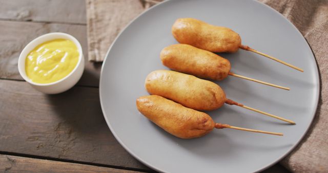 Four crispy corn dogs on skewers are laid out on a gray plate with a side of creamy mustard dip in a white bowl. This mouthwatering fast food snack is perfect for showcasing street food, casual dining, event catering, or recipe ideas for quick meals.