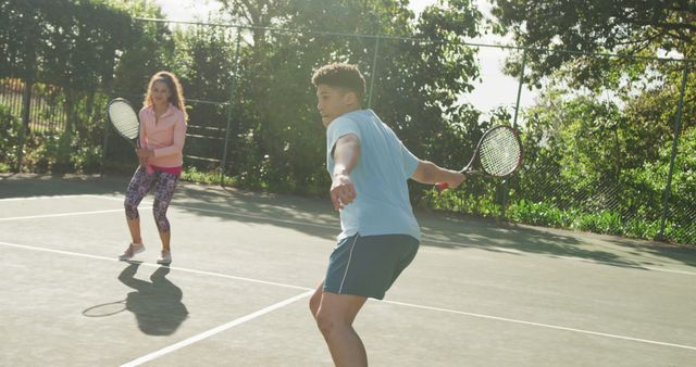 Two friends are playing tennis on an outdoor court. The scene depicts them actively engaging in the game on a sunny day. This stock image is perfect for use in advertising, sports magazines, fitness blogs, and social media campaigns promoting healthy lifestyles and recreational activities.