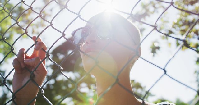 Person standing behind chain link fence, sunlight creating lens flare. Ideal for themes like freedom, contemplation, independence, or fashion. Suitable for use in blogs, magazines, or advertisements.