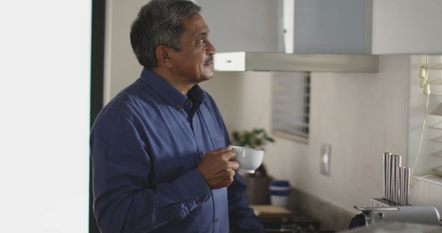 Elderly man standing in modern kitchen, holding coffee cup, seemingly lost in thought. Can be used for themes about relaxation, morning routine, mature lifestyle, or peaceful moments.