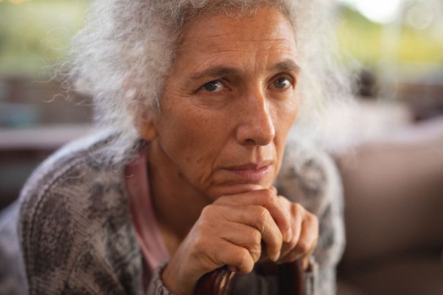 Elderly woman with grey hair leaning on a cane, looking thoughtful. Ideal for topics related to aging, retirement, senior lifestyle, and solitude. Can be used in articles, advertisements, or brochures about elderly care, mental health, and home living for seniors.