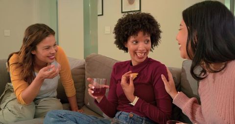 Diverse friends laughing on couch sharing wine and snacks during cozy home gathering