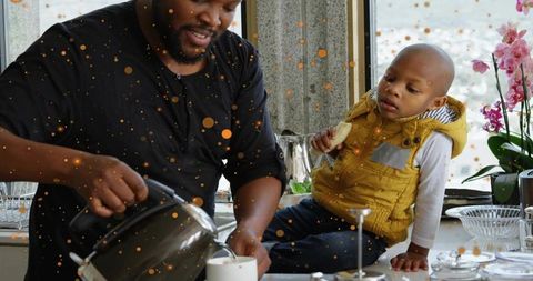 Father pouring hot drink for child on kitchen counter by window with orchids and daylight