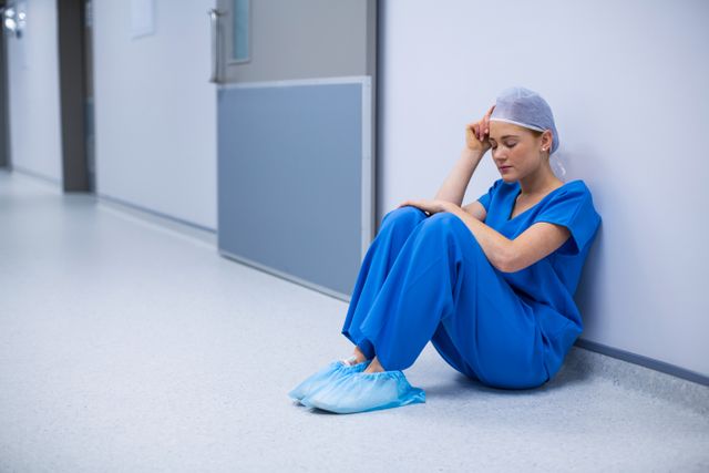 Nurse in blue scrubs sitting on floor in hospital corridor, appearing stressed and exhausted. Useful for illustrating healthcare worker stress, mental health in medical professions, and the demanding nature of healthcare jobs. Can be used in articles, blogs, and presentations about healthcare challenges, nurse burnout, and the importance of mental health support for medical staff.