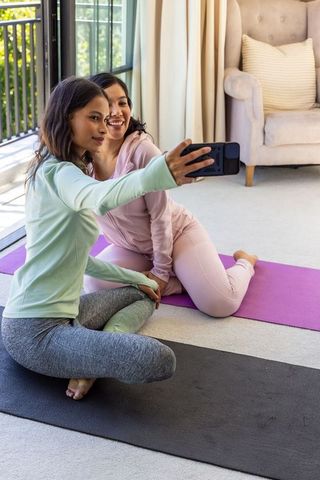 Diverse women taking selfie after home yoga session on sunlit balcony mats, smiling