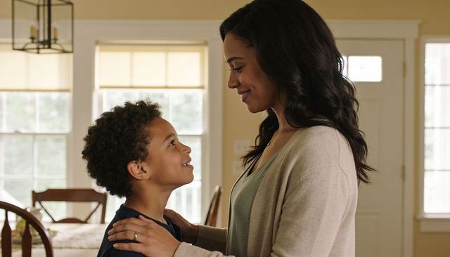Smiling mother holding son by shoulders in sunlit dining room showing wedding ring