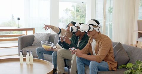 Diverse male friends playing multiplayer VR games on living room sofa wearing white headsets