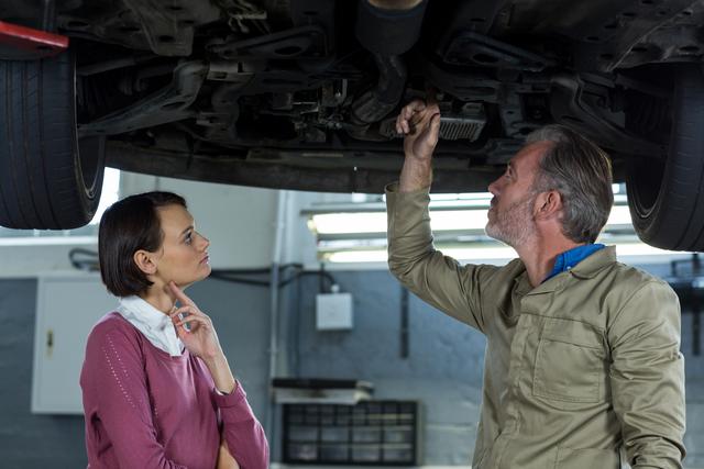 Mechanic explains car issues to customer under raised vehicle at repair shop. Ideal for automotive service advertisements, customer service training materials, auto repair tutorials, and car maintenance blog articles.