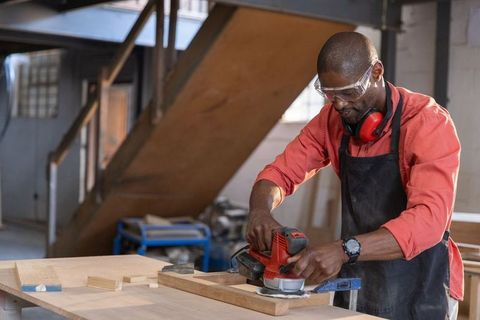 Senior african american craftsman sanding wooden plank in workshop with protective gear