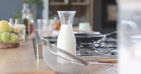 Glass carafe of milk on wooden counter with whisk, sieve and stove for breakfast baking