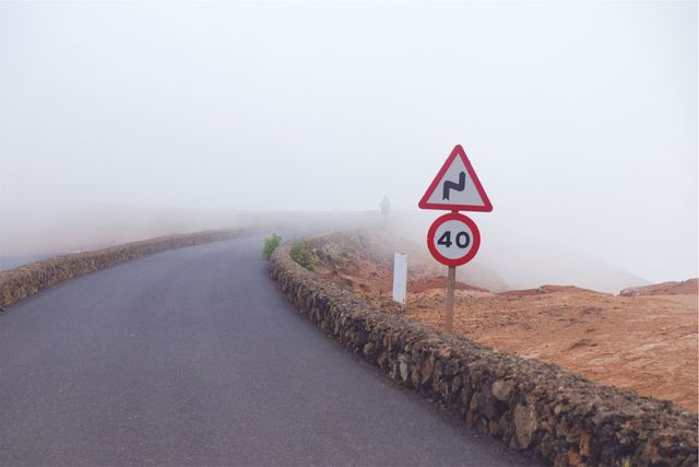 Curving narrow road disappears into thick mist with a warning sign displaying curves and 40 km/h speed limit. Ideal for themes of travel safety, weather conditions, rural driving, and scenic views.