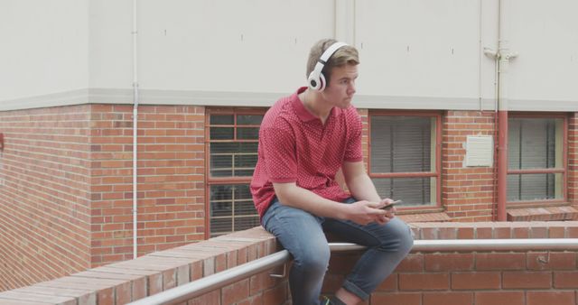 The young man is sitting on a building ledge, wearing headphones and holding a smartphone. His casual clothing includes a red shirt and jeans. This image captures a moment of relaxation and leisure in an urban setting. Suitable for use in campaigns or articles about youth culture, music, technology, and urban lifestyles.