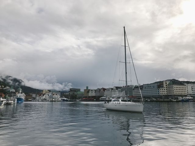 This scene shows a single sailing boat gliding through a peaceful harbor with a cloudy sky overhead. The reflection in the calm water enhances the serene atmosphere of a coastal town. This image is ideal for websites related to travel and tourism, nautical activities, and marine landscapes.