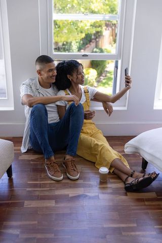 Young couple sitting on hardwood floor taking selfie by sunny window with coffee cup