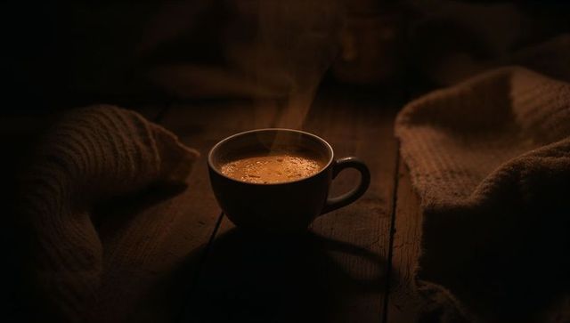 Steaming ceramic coffee cup on rustic wood table with cozy wool blankets and warm crema glow