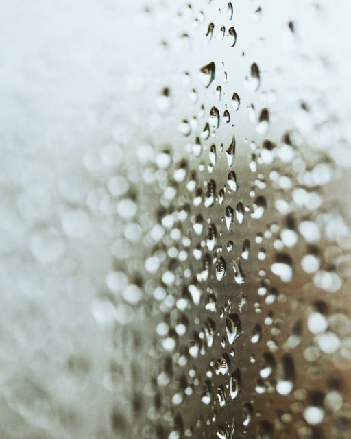 Raindrops are clinging to a window glass with a blurred background in the distance, creating a serene and calming atmosphere with the soft focus. This image can be used to convey themes of weather, rain, serenity, and tranquility. It is ideal for backgrounds, quotes about rain, or as a visual representation of rainy days and the peaceful moments that accompany them.