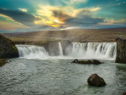 Sunset Glow Over Majestic Waterfall Cascading Into Wide River Across Rugged Highlands