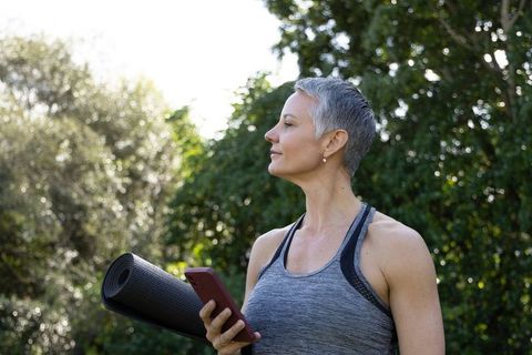 Mature woman holding yoga mat and checking phone while enjoying sunlight in park
