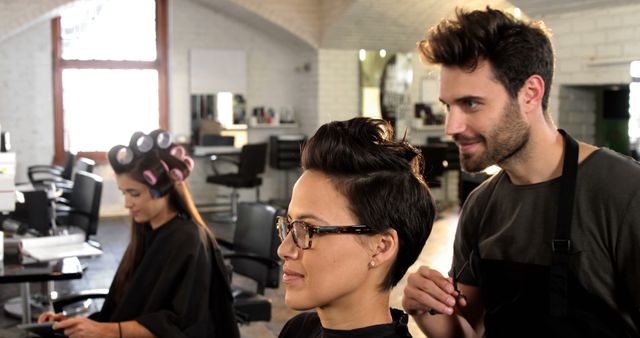 A young Asian woman gets a stylish haircut from a Caucasian male hairstylist in a modern salon, with copy space. Her reflection is visible in the mirror as another client sits in the background with rollers in her hair.