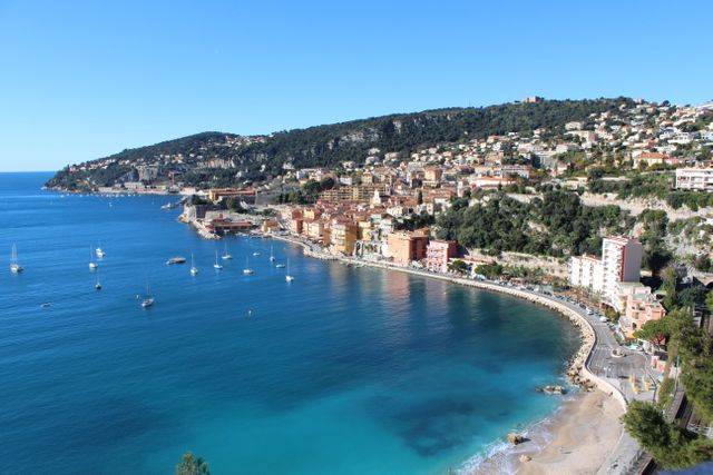 This image captures a stunning aerial view of a Mediterranean coastal town with boats anchored in clear blue water. The charming town features colorful buildings nestled on a hillside. This image is ideal for promoting travel, tourism, resort destinations, or vacation planning. It can also be used in brochures, travel websites, and advertisements related to beautiful coastal locations.