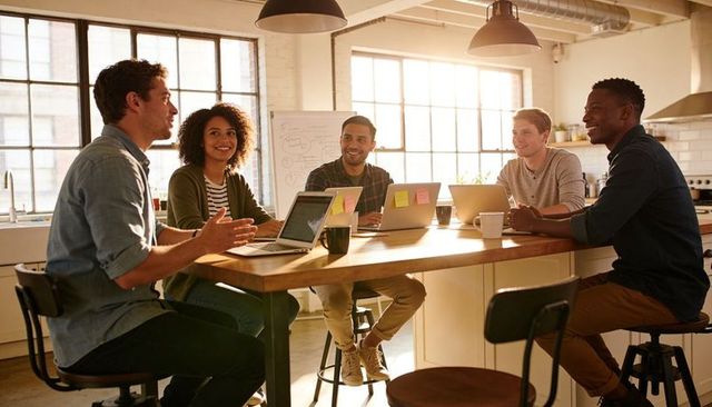 Young diverse startup team brainstorming around kitchen island with laptops, coffee, sunlight