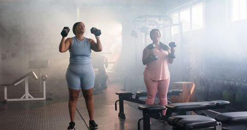 Diverse female friends lifting dumbbells during gym strength training in bright activewear