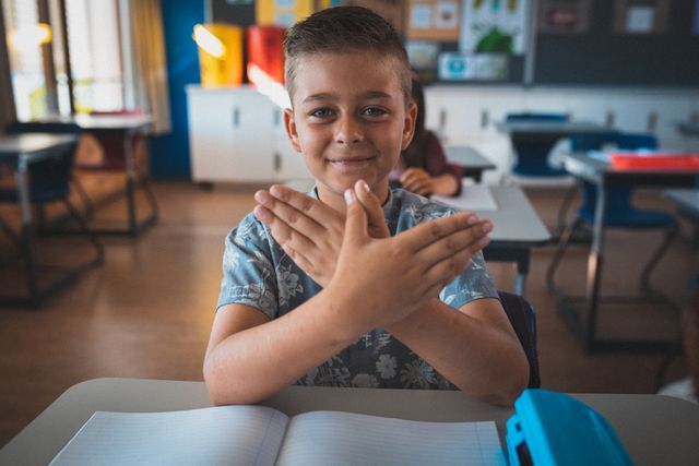 Young boy sitting at desk in classroom, smiling and making bird shape with hands. Ideal for educational content, school promotions, childhood development articles, and creative learning resources.
