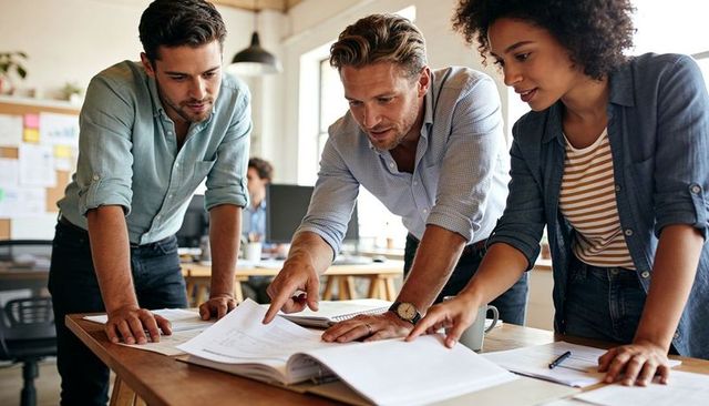 Collaborating team reviewing plans at desk, pointing at binder in bright office