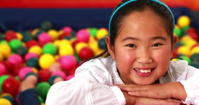 Smiling girl in a white shirt laying in colorful ball pit. Suitable for themes related to childhood joy, playground fun, indoor children's activities, and family entertainment. Ideal for use in advertisements for play centers, editorial content on kids' playtime, or educational materials focusing on developmental play.