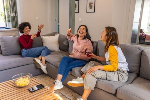 Diverse women laughing and chatting on sectional sofa, enjoying drinks and snacks