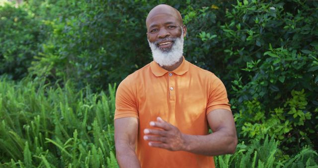 A smiling man with a beard, wearing an orange polo shirt, enjoying a beautiful day outdoors surrounded by lush, green plants. Ideal for promoting outdoor activities, healthy living, and senior well-being. Suitable for advertisements, lifestyle blogs, and websites related to nature, fashion, and wellness.