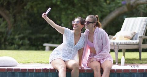 Two young women sitting on pool edge taking selfie while enjoying sunny backyard afternoon