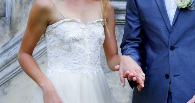 A bride in a lace wedding dress and a groom in a blue suit are holding hands, symbolizing their union and commitment. Their wedding attire and the act of holding hands capture a moment of love and partnership on their special day.
