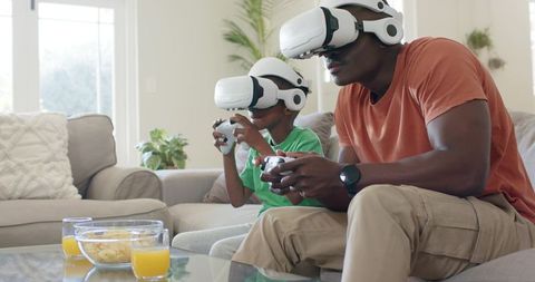 African American Father and Son Playing Virtual Reality Games Together at Home on Sofa