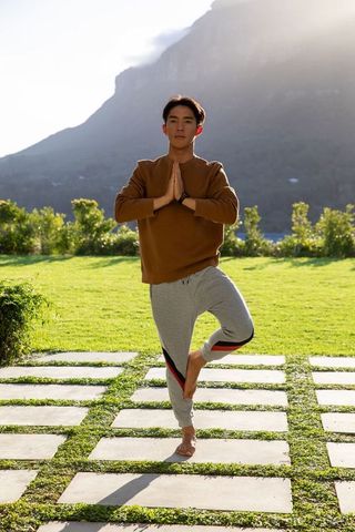 Asian man practicing tree pose on stone pavers near mountain shoreline barefoot smartwatch