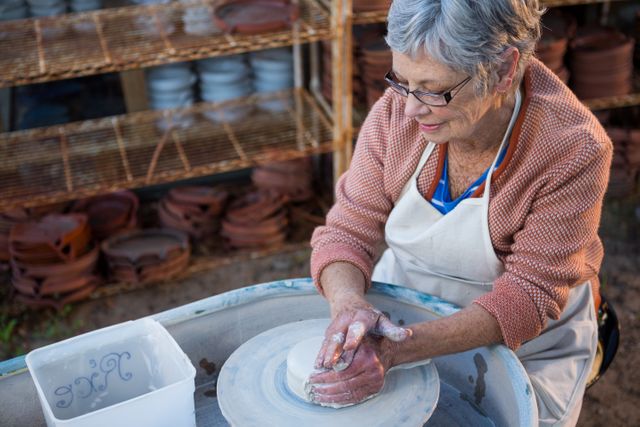 Senior woman shaping clay on potter's wheel in pottery workshop. Ideal for illustrating creativity, craftsmanship, and hobbies among elderly individuals. Useful for articles on traditional arts, senior activities, and handmade crafts.
