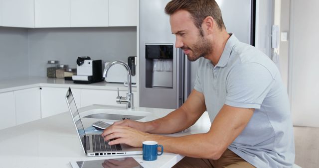 A man working on a laptop at a kitchen counter, focusing on tasks. This can be used for topics related to remote work, home office setups, productivity tips, and technology in daily life. The image shows a modern, clean kitchen environment.