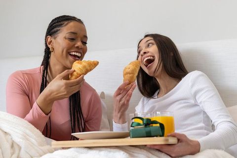 Sharing breakfast in bed with croissants and orange juice, laughing friends enjoying morning