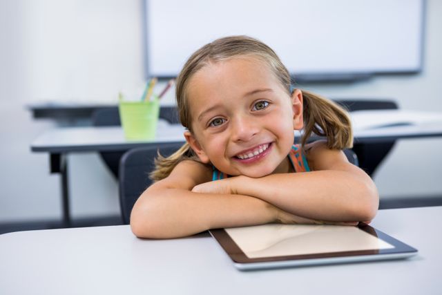 Young girl with pigtails smiling and leaning on a digital tablet in a classroom. Ideal for educational content, technology in education, school advertisements, and child development articles.