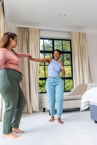 Two african american women dancing and laughing in bright bedroom holding hands