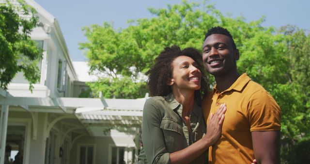 An African American couple is embracing and smiling outdoors on a sunny day, standing by a house with greenery and trees in the background. Useful for topics on love, relationships, summer activities, home life, outdoor enjoyment, and happiness. Perfect for lifestyle blogs, relationship counseling, and family-oriented promotions.
